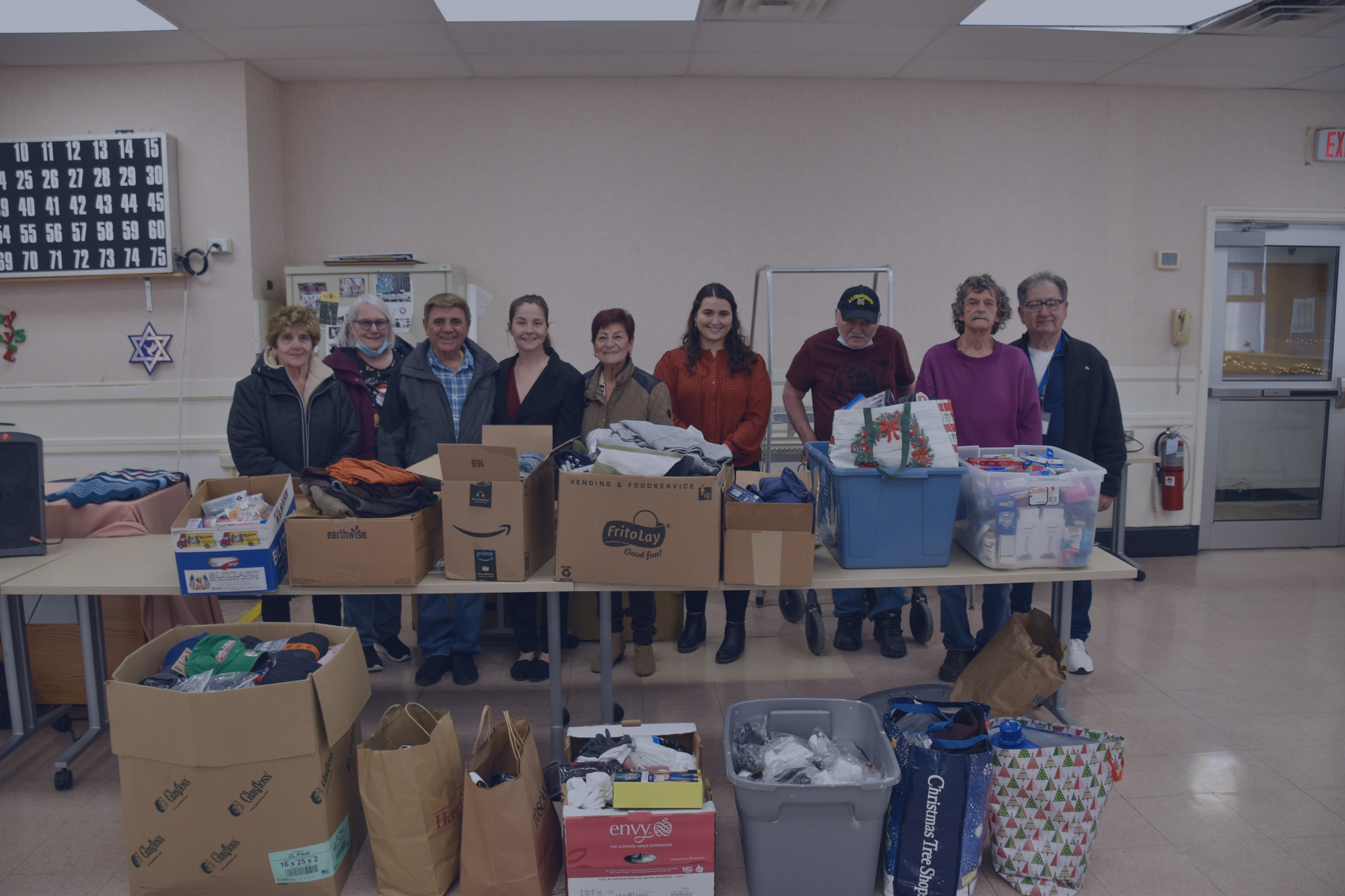 Community members pose behind tables covered with boxes and bins of donated socks, clothing, hygiene supplies, and household items collected for the City of Everett’s Veterans Comfort Drive in a community center.