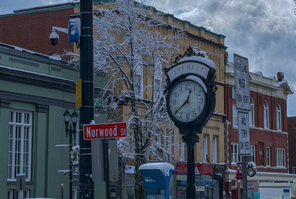 Snow-covered Everett street scene featuring the historic Everett clock at the corner of Norwood Street, with brick buildings and winter trees in the background.