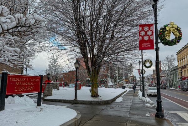 Snow-covered courtyard outside the Parlin Memorial Library, with trees, holiday wreaths on street lamps and a wet sidewalk along Broadway in Everett.