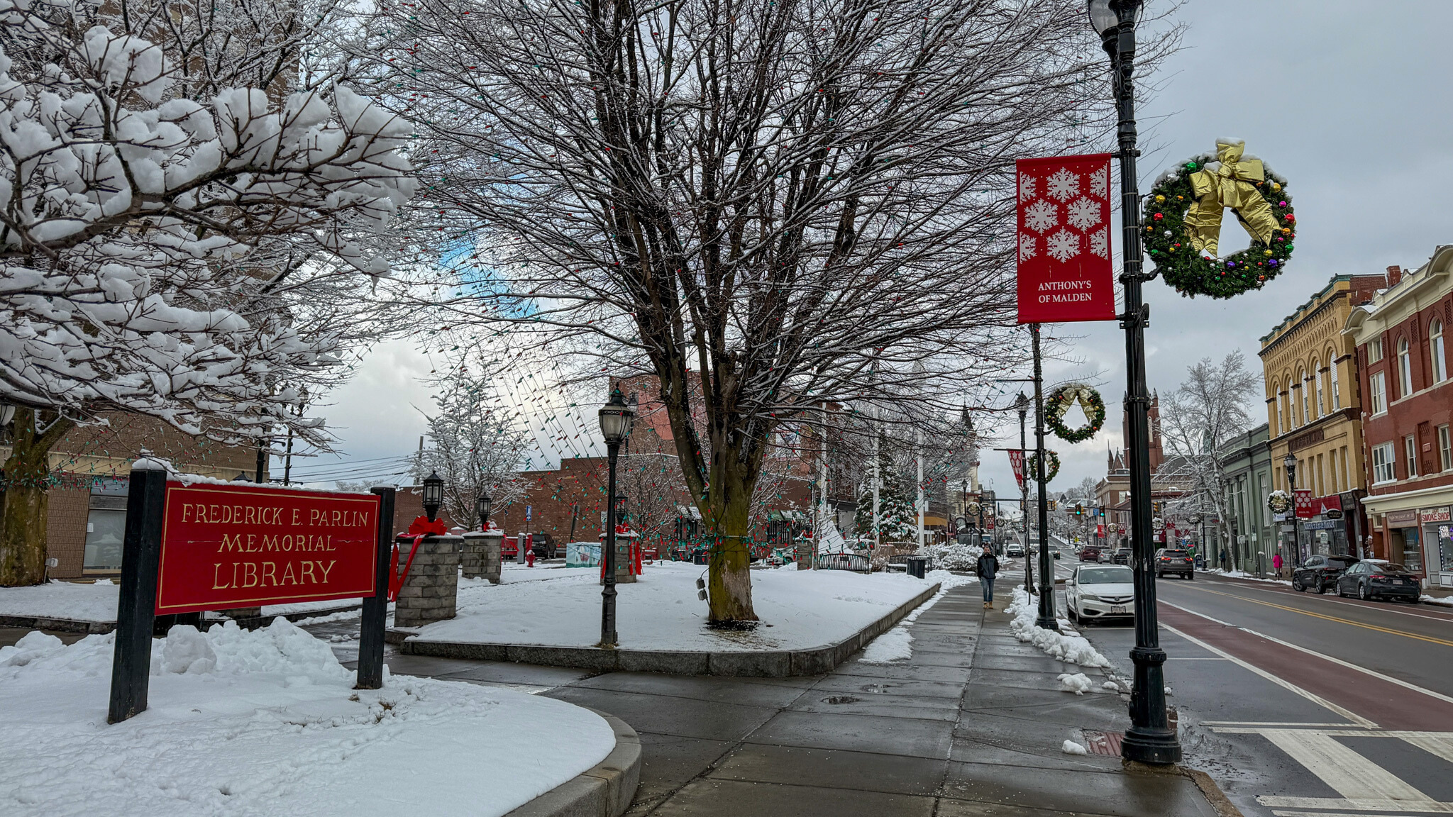 Snow-covered courtyard outside the Parlin Memorial Library, with trees, holiday wreaths on street lamps and a wet sidewalk along Broadway in Everett.