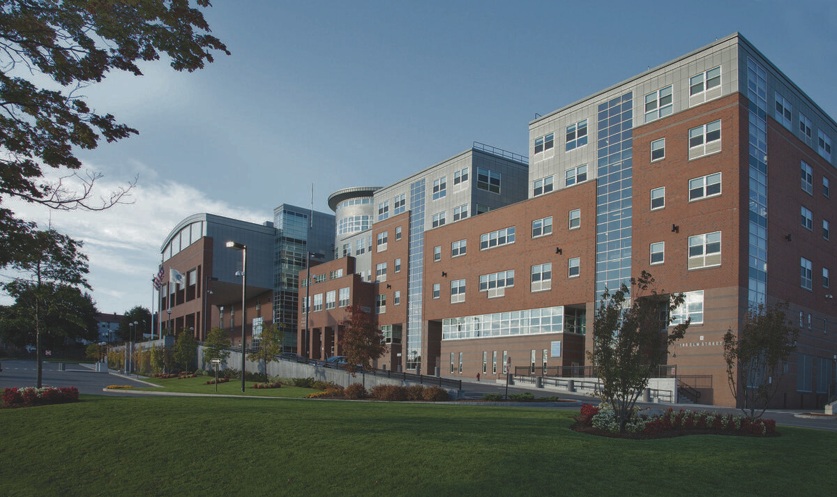 Outdoor photo of Everett High School. A landscaped green lawn with trees, flower beds, and a paved walkway sits in the foreground, with flagpoles and streetlights visible near the entrance under a clear blue sky.
