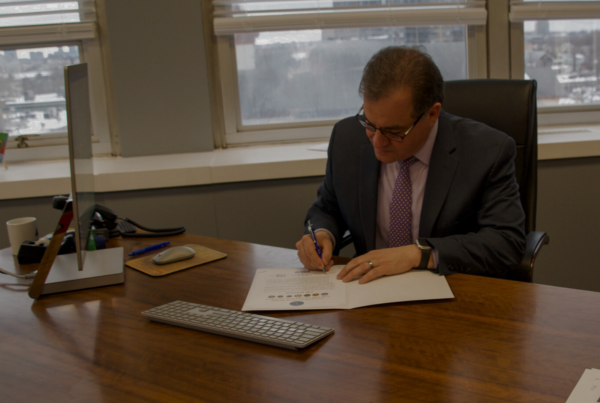 Mayor Robert Van Campen sits behind a desk in his office while holding a pen and looking down as he signs the Statement of Support for the Employer Support for the Guard and Reserve.
