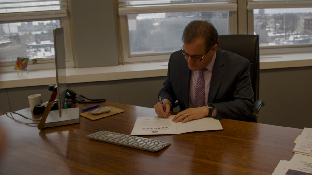 Mayor Robert Van Campen sits behind a desk in his office while holding a pen and looking down as he signs the Statement of Support for the Employer Support for the Guard and Reserve.