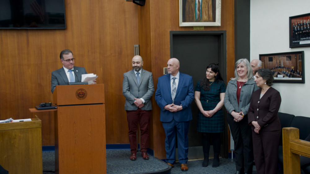 Mayor Robert Van Campen stands at a podium inside the Everett City Council Chamber as he reads from a piece of paper. Standing alongside him is his senior staff that he is introducing to the audience.