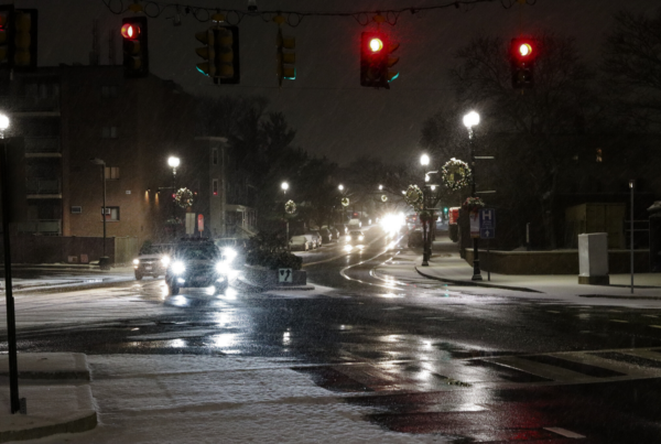 Snow falls lightly over a city intersection at night, where red traffic lights glow above wet, reflective pavement. Cars with bright headlights drive along the slick road, and holiday wreaths hang from lampposts lining the snowy sidewalks.