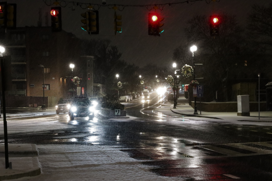 Snow falls lightly over a city intersection at night, where red traffic lights glow above wet, reflective pavement. Cars with bright headlights drive along the slick road, and holiday wreaths hang from lampposts lining the snowy sidewalks.