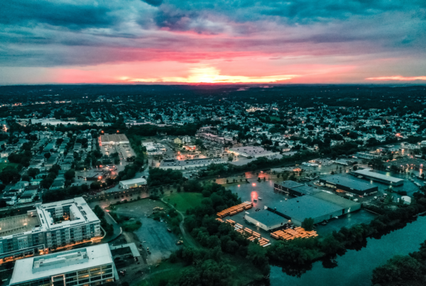 Aerial view of Everett at sunset, showing a dense neighborhood of homes, commercial buildings, and roads stretching toward the horizon. The sky glows with pink and orange hues beneath dark clouds, while streetlights and parking lots begin to illuminate the city below.