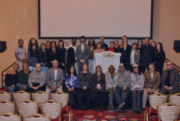 Large group of adults who received the last round of funding pose for a group photo on stage in a banquet hall at Encore casino. A large projection screen hangs behind them, with speakers on both sides of the stage and rows of empty chairs visible in the foreground.