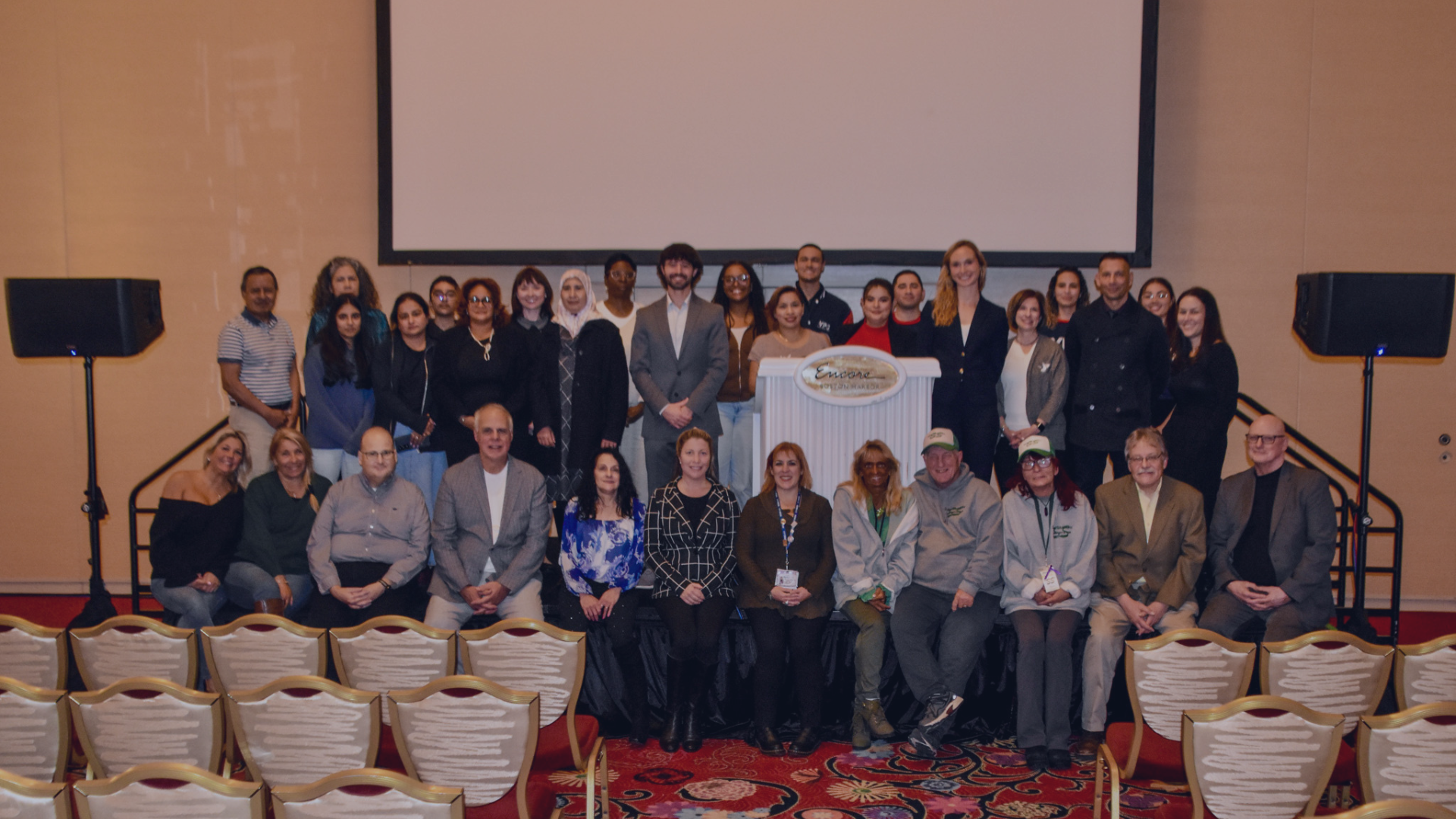 Large group of adults who received the last round of funding pose for a group photo on stage in a banquet hall at Encore casino. A large projection screen hangs behind them, with speakers on both sides of the stage and rows of empty chairs visible in the foreground.
