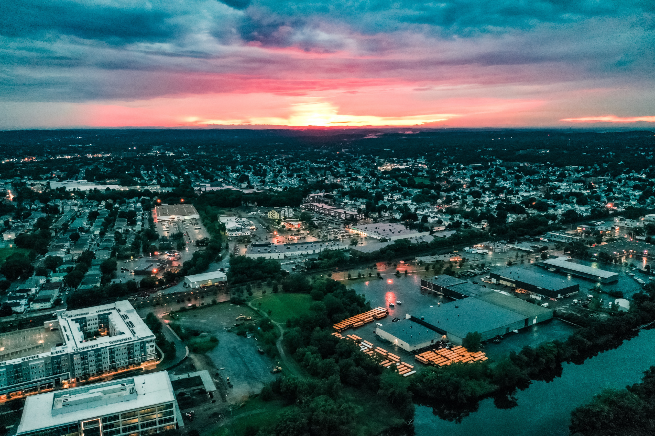 Aerial view of Everett at sunset, showing a dense neighborhood of homes, commercial buildings, and roads stretching toward the horizon. The sky glows with pink and orange hues beneath dark clouds, while streetlights and parking lots begin to illuminate the city below.