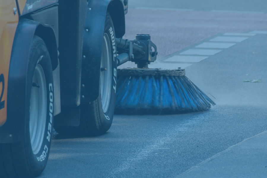 Close-up of a municipal street sweeper using a rotating blue brush to clean debris from the roadway near the curb, with water spraying onto the pavement during the sweeping process.