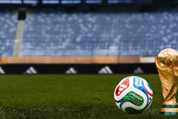 A FIFA World Cup trophy and a soccer ball rest on a grass field inside a stadium, with empty blue seats blurred in the background.