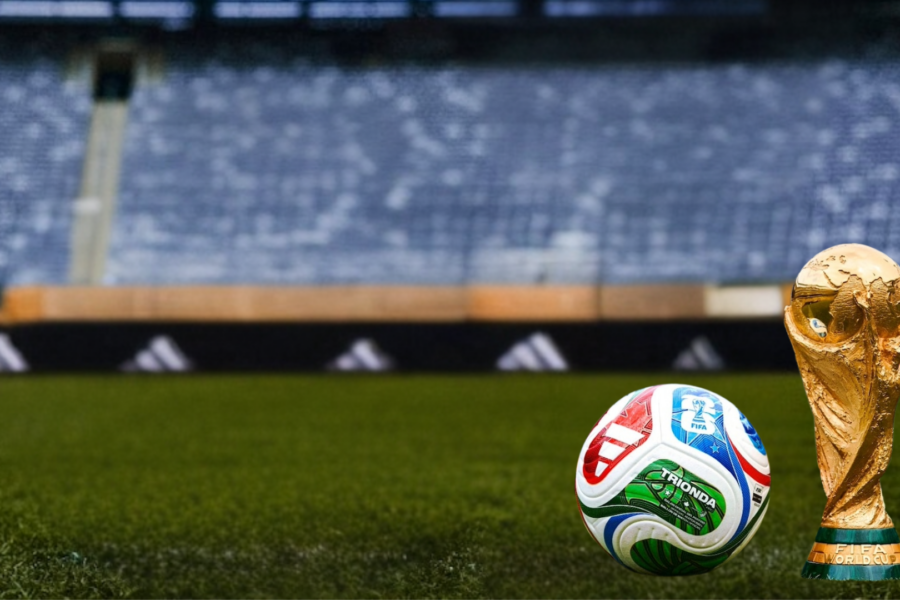 A FIFA World Cup trophy and a soccer ball rest on a grass field inside a stadium, with empty blue seats blurred in the background.