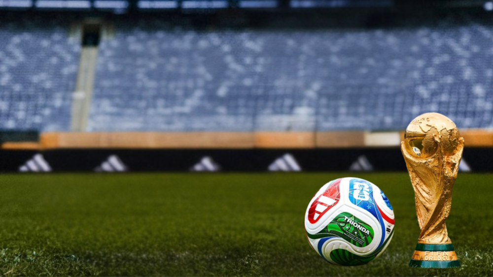 A FIFA World Cup trophy and a soccer ball rest on a grass field inside a stadium, with empty blue seats blurred in the background.