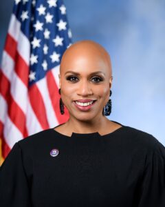 Congresswoman Pressley smiling in a black top and earrings with an American flag in the background.