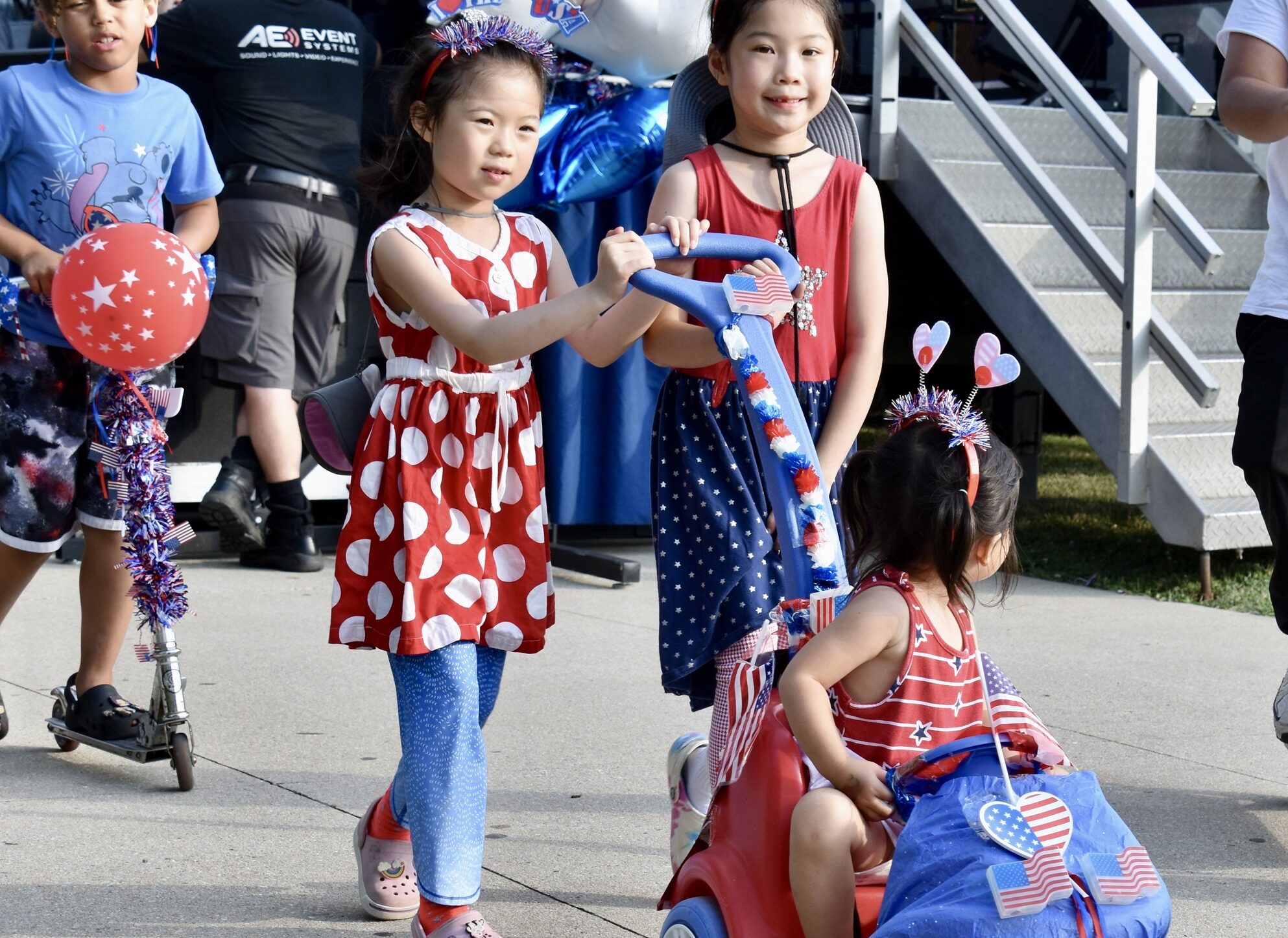 Two girls in red, white, and blue garb push their little sister in a decorated wagon.