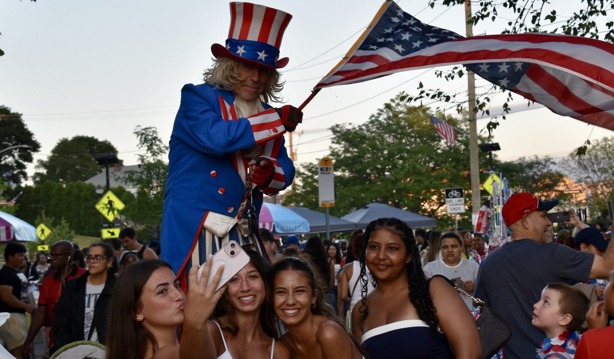 Four girls take a selfie with Uncle Sam on stilts, who waves the American flag.