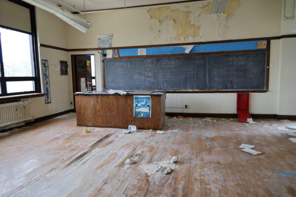 An empty classroom with a chalkboard and wooden desk. The floors and walls are deteriorated