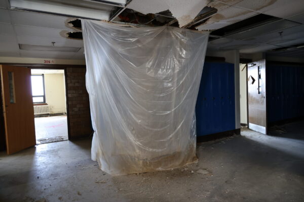 A tarp hangs from the ceiling to floor in the school hallway showing signs of water damage.