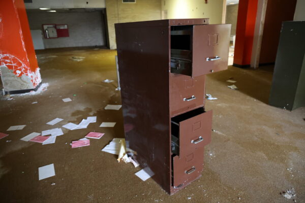A file cabinet in the middle of an old deteriorated library with the draws half open and papers on the ground next to it