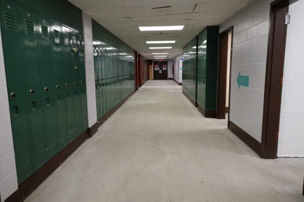 Empty school hallway with green lockers showing the tiles have been removed all that's left is concrete on the floor.