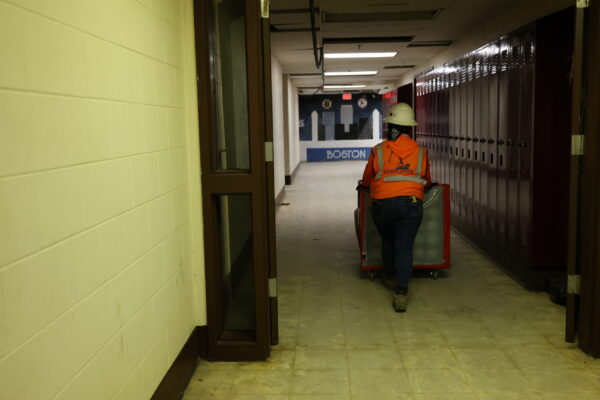 A woman in an orange vest and hard hat seen pushing a crate down an empty school hallway