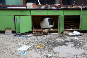 An old science lab in disarray, green cabinet doors are open showing an old computer monitor from the early 2000s with broken glass and papers strewn about the classrom